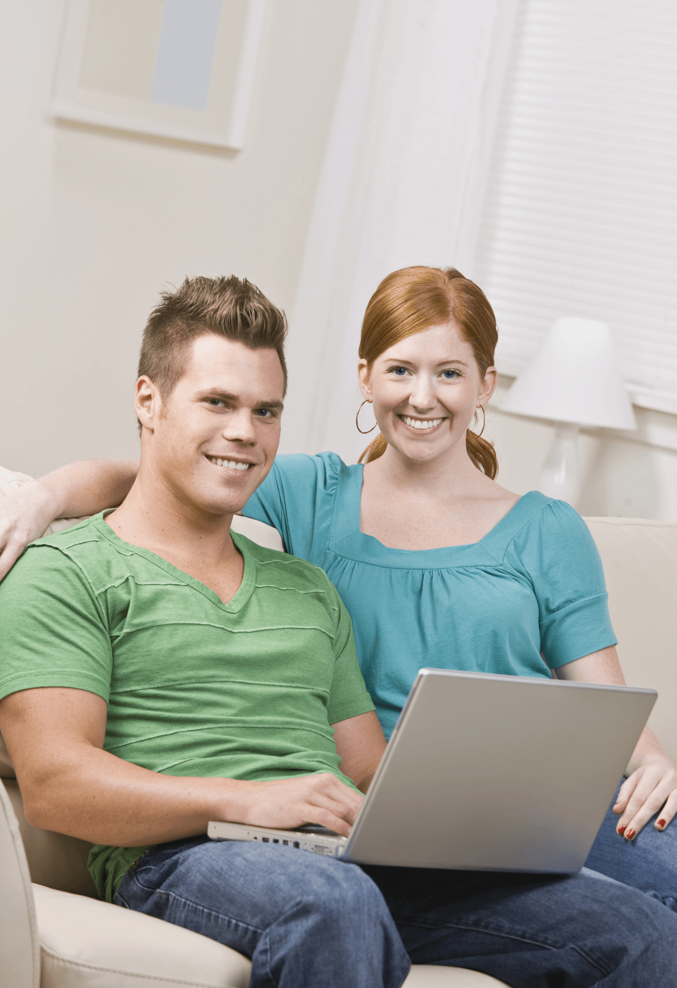 A smiling couple sitting together on a couch, both wearing casual clothes. The man is using a laptop while the woman sits next to him, offering support. They look comfortable and engaged, possibly participating in online counseling from home.