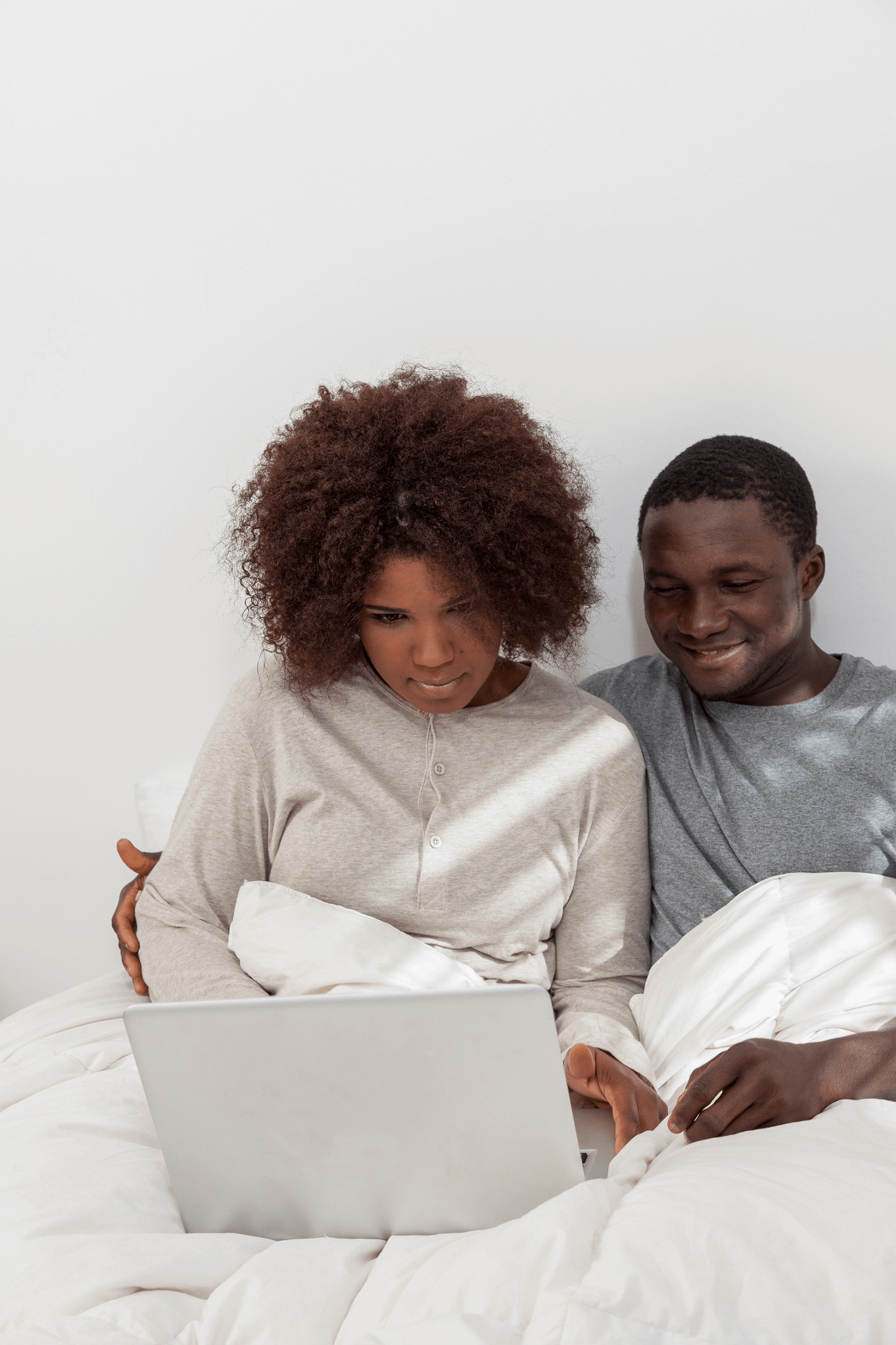 A couple sitting together in bed, the woman focused on a laptop while the man smiles at the screen. Both are dressed in comfortable loungewear, possibly engaged in an online Christian counseling session as they lean on each other for support.