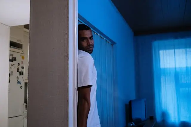 A man in a white t-shirt leans against a doorframe, partially hidden in the shadows. His facial expression appears tense or suspicious as he looks toward the camera. The room behind him is dimly lit with a cool blue hue and he is pondering the_patterns_of_paranoia_the_fear_of_loosing_control